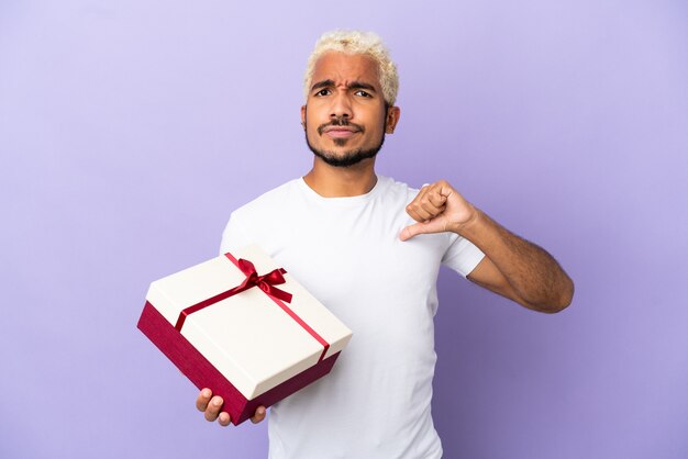 Young Colombian man holding a gift isolated on purple background proud and self-satisfied