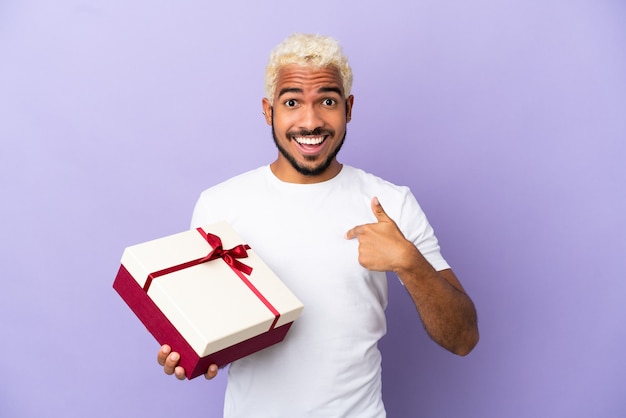 Young Colombian man holding a gift isolated on purple background with surprise facial expression