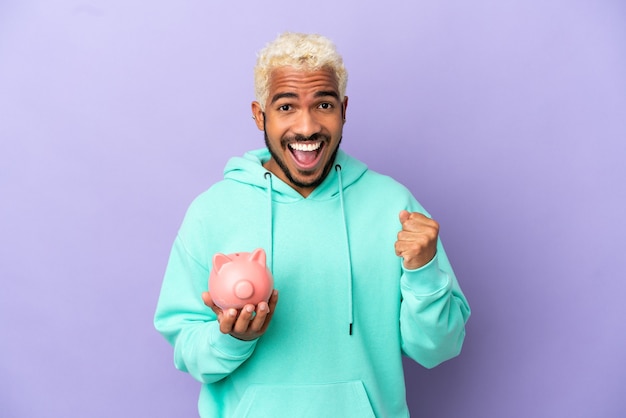 Young Colombian man holding a piggybank isolated on purple background celebrating a victory in winner position