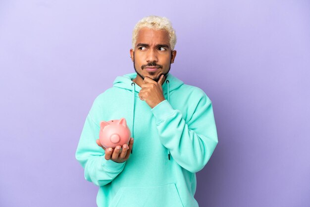 Young Colombian man holding a piggybank isolated on purple background having doubts and thinking