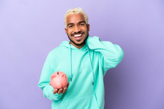 Young Colombian man holding a piggybank isolated on purple background laughing