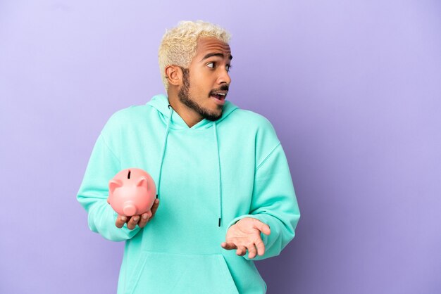 Young Colombian man holding a piggybank isolated on purple background with surprise expression while looking side