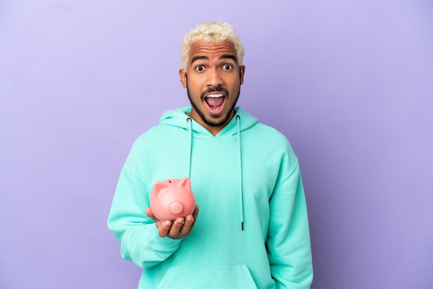 Young Colombian man holding a piggybank isolated on purple background with surprise facial expression