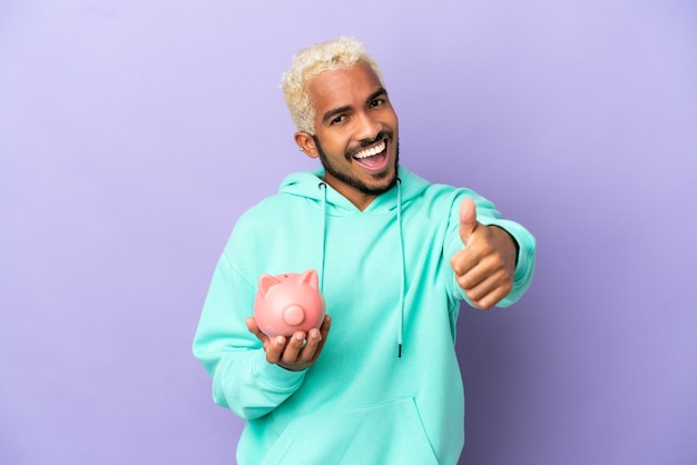 Young Colombian man holding a piggybank isolated on purple background with thumbs up because something good has happened