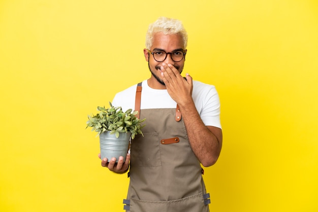 Young Colombian man holding a plant isolated on yellow background happy and smiling covering mouth with hand