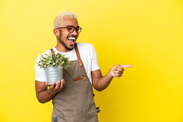 Young Colombian man holding a plant isolated on yellow background pointing finger to the side and presenting a product