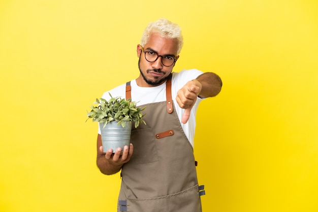 Young Colombian man holding a plant isolated on yellow background showing thumb down with negative expression