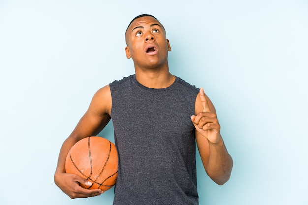 Young colombian man playing basketball pointing upside with opened mouth.