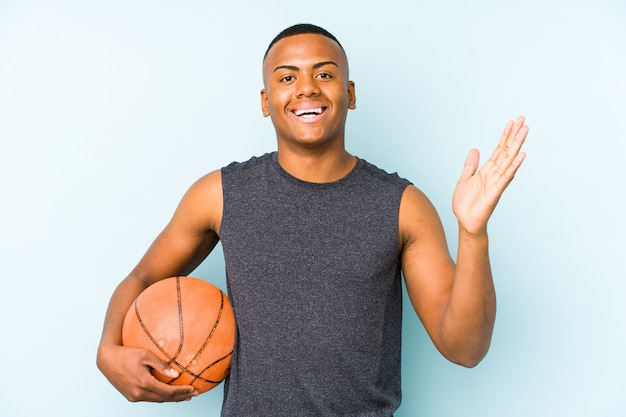 Young colombian man playing basketball receiving a pleasant surprise, excited and raising hands.