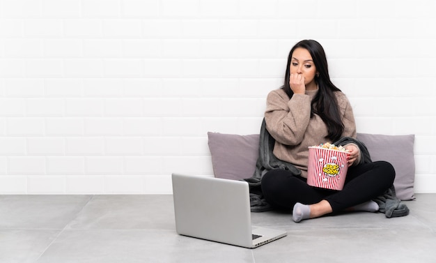 Young Colombian woman holding a bowl of popcorns and showing a film in a laptop having doubts