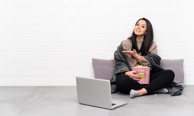 Young Colombian woman holding a bowl of popcorns and showing a film in a laptop presenting an idea while looking smiling towards