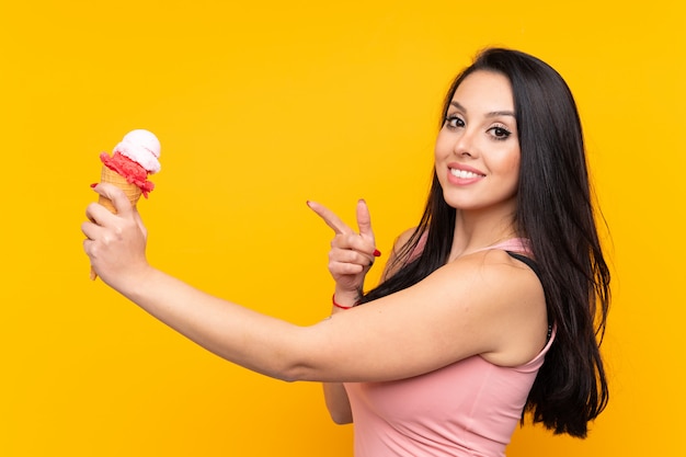 孤立した黄色の壁にコルネットアイスクリームを押しながらそれを指している若いコロンビアの女性