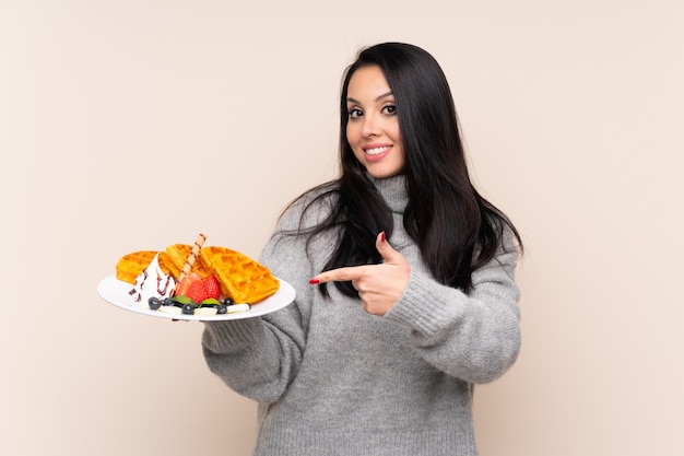 Young Colombian woman holding waffles over isolated wall and pointing it