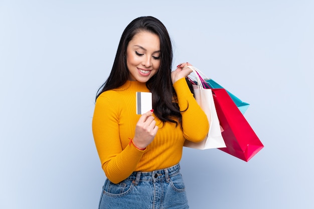 Young Colombian woman over isolated blue wall holding shopping bags and a credit card