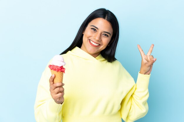 笑みを浮かべて勝利のサインを示す青い壁にコルネットアイスクリームを持つ若いコロンビア女性