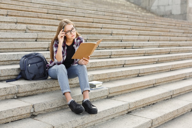 Young concentrated female student making notes in notebook, sitting on university stairs. Education, inspiration and remote working concept.