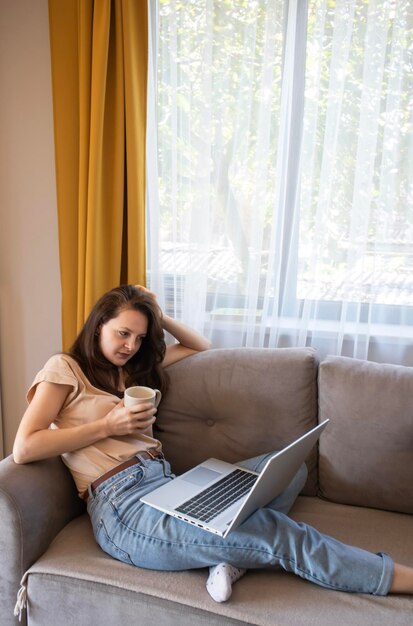 A young concentrated woman is drinking a cup of coffee during working from home A girl is thinking and sitting on the sofa with a laptop