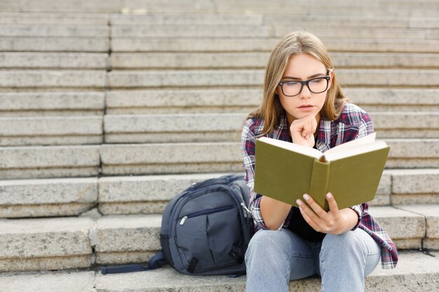 Young concentrated woman reading book on university stairs, preparing for exams at college. Education concept, copy space