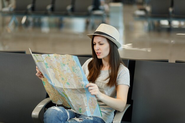 Young concerned traveler tourist woman holding paper map, searching route, wait in lobby hall at international airport