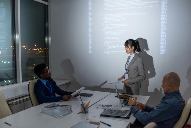 Young confident Asian businesswoman with papers interacting with African male colleague sitting by desk in front of her