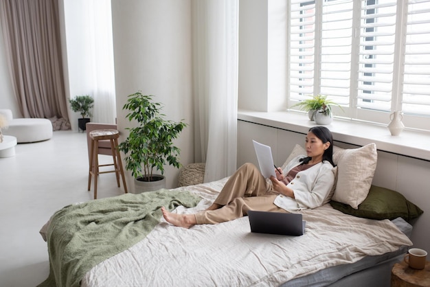 Молодая уверенная в себе азиатка в повседневной одежде сидит на кровати и читает газеты