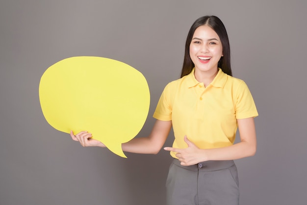 Young confident beautiful woman wearing yellow shirt is holding a yellow empty speech on grey studio