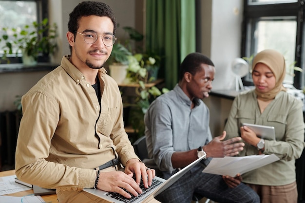 Young confident broker looking at camera while typing on laptop keyboard