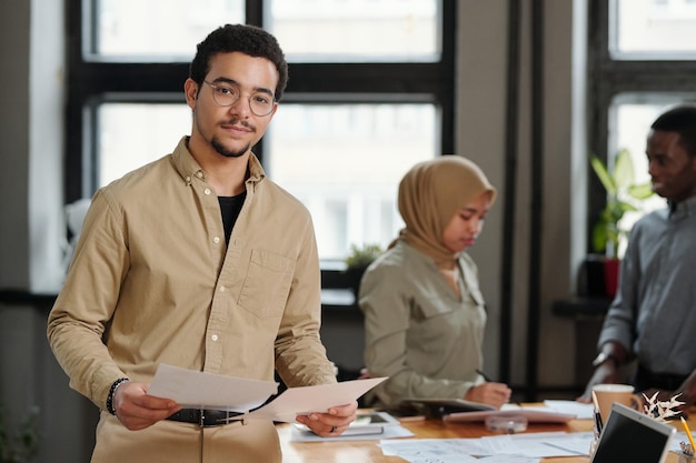 Young confident broker with financial documents standing in front of camera