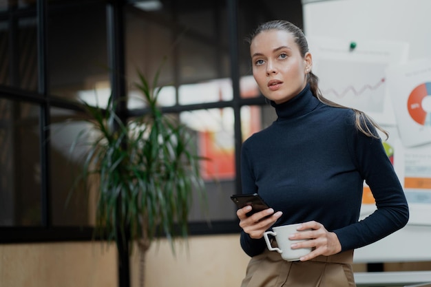 Young confident brownhaired woman holding a phone and a cup of coffee blue jacket