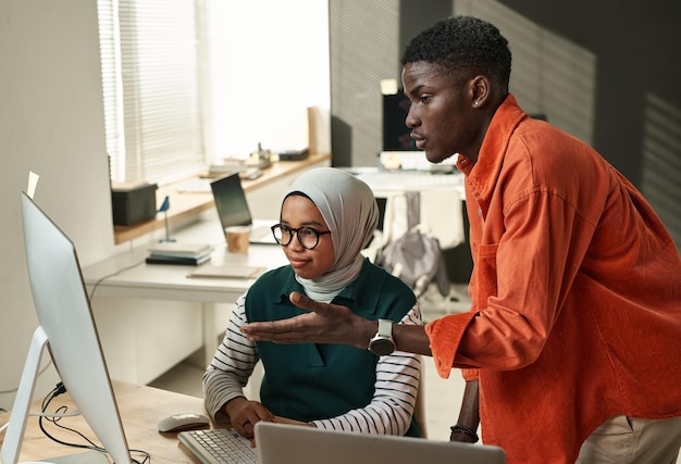 Young confident businessman consulting with muslim female\
colleague during presentation of online in