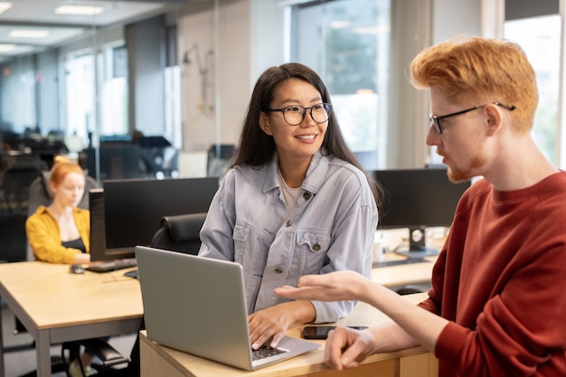 Young confident businessman in eyeglasses and casualwear making presentation for his female colleague in front of laptop at meeting