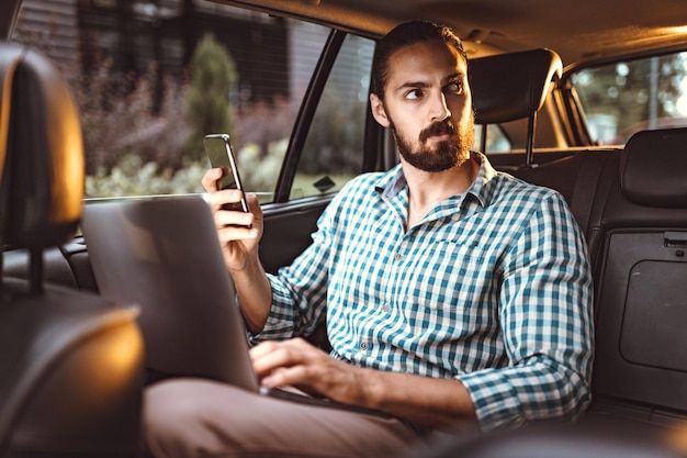 若い自信のあるビジネスマンは、スマートフォンを持って、車の後部座席でラップトップで話し、作業しています。