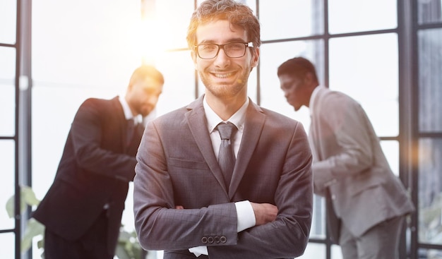 Young Confident Businessman Smiling at the Camera with Arm Crossing Over his Chest Against Coworkers Background