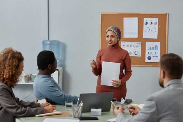 Young confident businesswoman in headscarf standing in front of colleagues