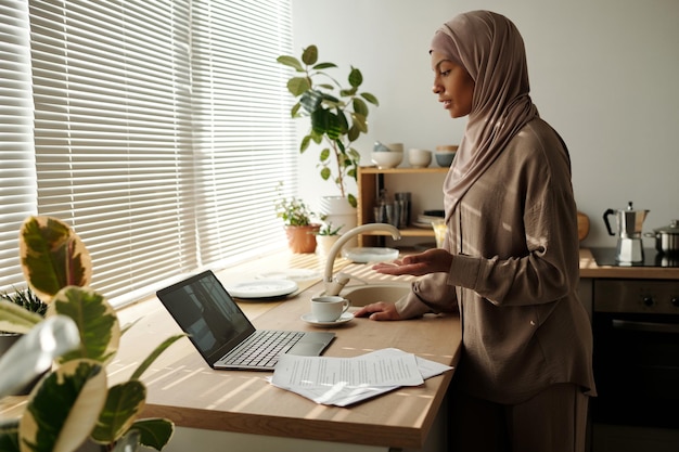 히자브를 입은 젊은 자신감 있는 사업가 여성이 동료와 대화하는 동안 노트북 화면을 보고 있습니다.