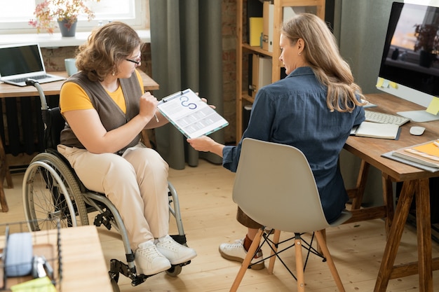 Photo young confident businesswoman in wheelchair showing financial document with graph and charts to colleague during presentation