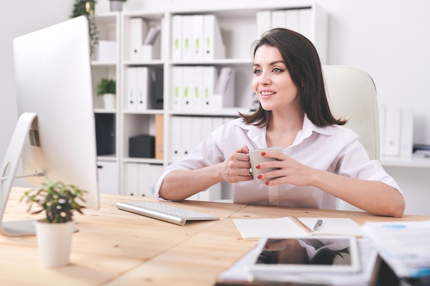 Young confident businesswoman with mug sitting by desk in front of computer screen, having tea and talking through video chat