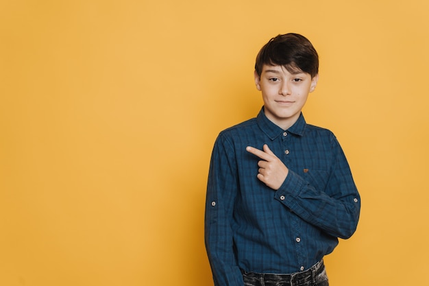 Young confident casual dressed boy pointing at copy space, standing against yellow background. Success people concept