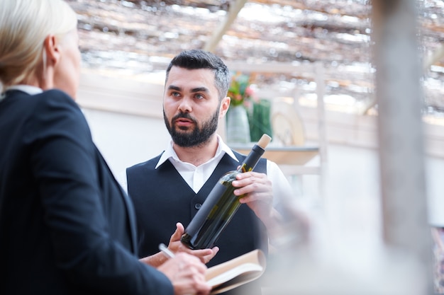 Foto giovane cavista fiducioso con una bottiglia di vino che parla con il suo collega durante la discussione delle sue caratteristiche al lavoro