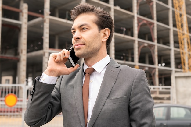 Young confident contractor in elegant suit standing on building site while phoning one of partners before meeting