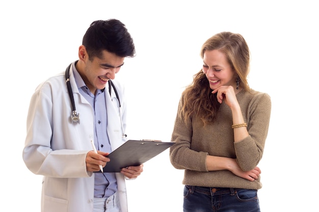 Молодой уверенный в себе врач в белом халате со стетоскопом смотрит на бумаги с молодой милой женщиной