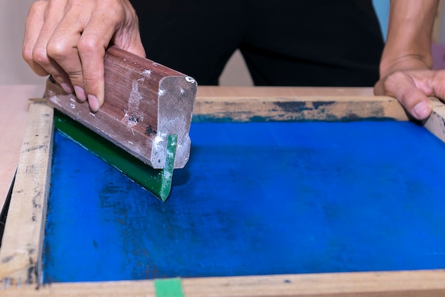 Young confident experienced male working with squeegee on a small factoryworker man using printmaking toolsscreen printing on clothing fabric technique