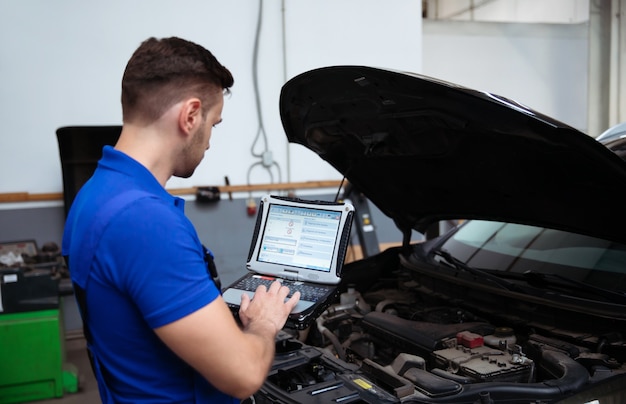 Il giovane meccanico fiducioso e bello con il laptop in mano conduce la diagnostica del computer per gli errori nei sistemi dell'auto