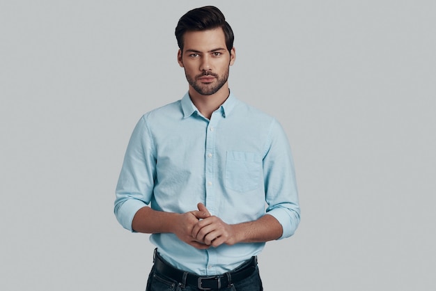 Young and confident. Handsome young man looking at camera and keeping hands clasped while standing against grey background