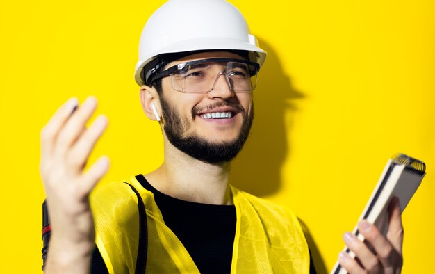 young confident man, builder engineer holding notebook, using wireless earphones, wearing safety helmet and goggles for construction on yellow wall.