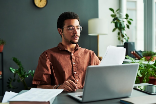 Young confident man with papers sitting in front of laptop