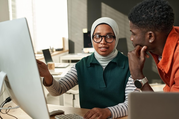 Young confident muslim woman in hijab pointing at computer\
screen and looking at african american ma