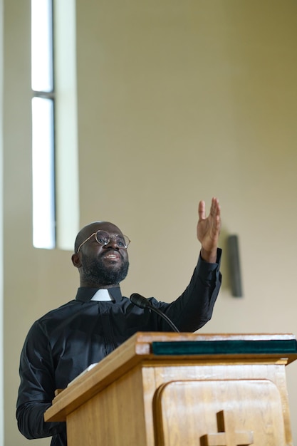 Young confident priest in black shirt with clerical collar standing by pulpit
