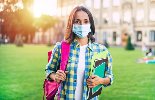 Young confident student woman in protect the medical mask with books and documents in hands and standing on university campus 