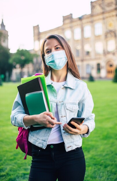 Young confident student woman in protect the medical mask with books and documents in hands and standing on university campus 